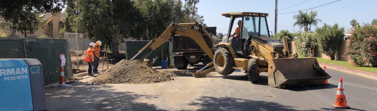 Two workers directing an excavator digging a trench in the street.